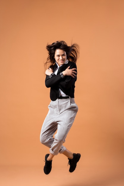 Felice ragazza dai capelli scuri vestita con camicia bianca, pantaloni grigi, giacca nera e scarpe da ginnastica nere salta sullo sfondo beige in studio.