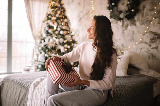 Felice ragazza dai capelli scuri vestita con maglione bianco e pantaloni tiene un regalo di Capodanno