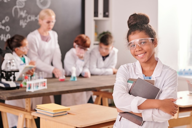 Felice ragazza della scuola secondaria di razza mista in camice bianco che ti guarda con un sorriso a trentadue denti contro i compagni di classe e l'insegnante durante il lavoro di laboratorio