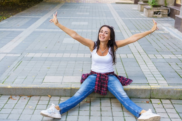 Felice ragazza in maglietta bianca blue jeans e scarpe da ginnastica bianche si siede sul marciapiede nel parco durante il giorno che...