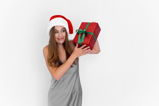 Felice ragazza positiva in cappello di Babbo Natale che scuote la confezione regalo mentre si trova su sfondo bianco celebrazione