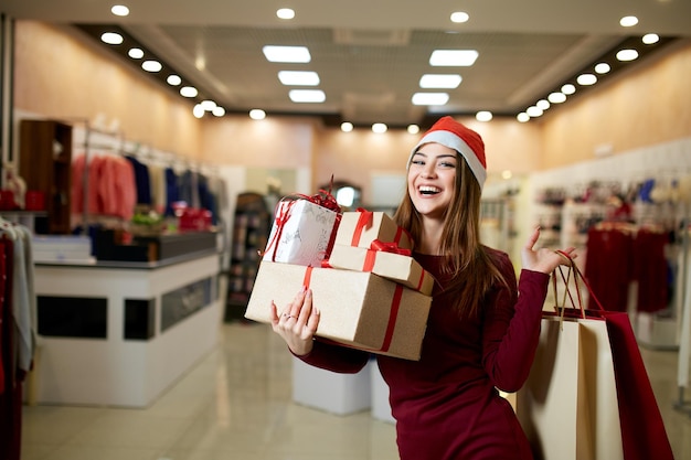 Felice ragazza shopping regali nel centro commerciale in vendita di Natale Capodanno vacanze shopping idea concetto Donna sorridente con carta colorata presenta borse e scatole regalo che indossano cappello di Natale in negozio o negozio