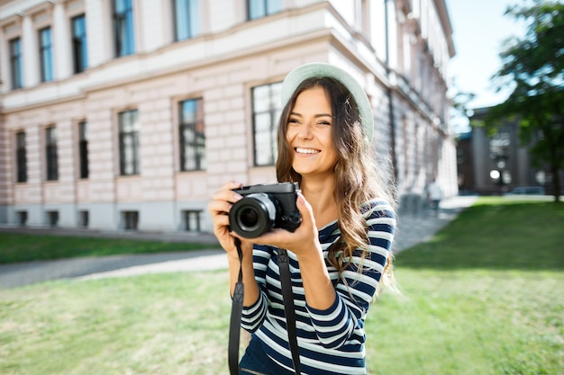 Felice ragazza turistica con capelli ricci che indossa un cappello, che tiene la macchina fotografica e sorridente alla vecchia città europea, viaggiando.