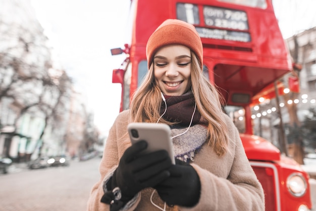 Felice ragazza vestita con abiti caldi e un guanto si trova in strada con uno smartphone in mano, ascolta la musica in cuffia e sorride