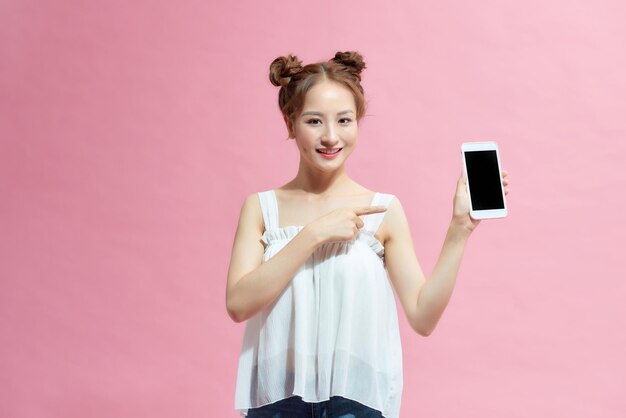 Felice ragazza vestita d'estate che punta il dito sul telefono cellulare con schermo vuoto isolato su sfondo rosa