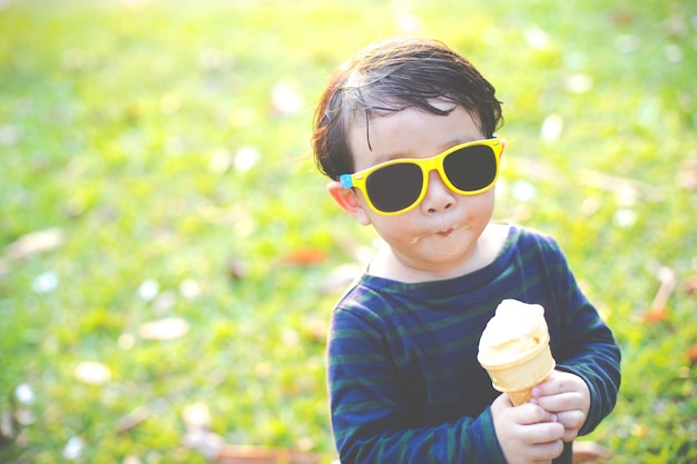 Felice ragazzino asiatico che mangia un cono gelato con occhiali da sole in giardino, soft focus