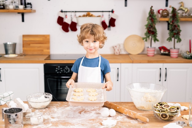 Felice ragazzo carino con biscotti fatti in casa crudi ti guarda mentre è in piedi dal tavolo della cucina