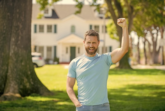 Felice ragazzo con la barba lunga in piedi vicino alla nuova casa che celebra il quartiere di successo