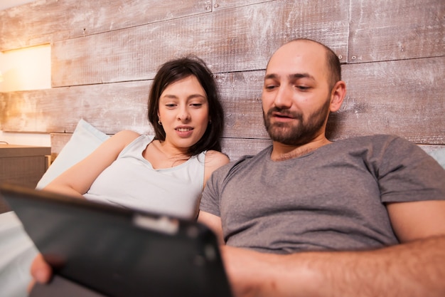 Felice ragazzo e ragazza che indossano il pigiama sdraiato a letto navigando sul computer tablet.