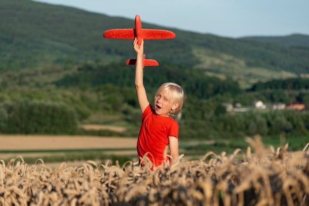 Felice ragazzo in maglietta rossa gioca con l'aereo giocattolo nel campo di grano sullo sfondo delle montagne Voli e viaggi con i bambini