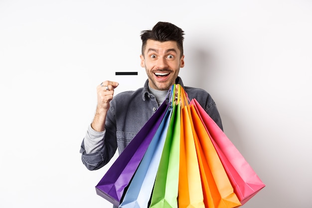 Felice ragazzo shopaholic tenendo colorate borse della spesa e mostrando la carta di credito in plastica, sorridendo eccitato, in piedi sfondo bianco.