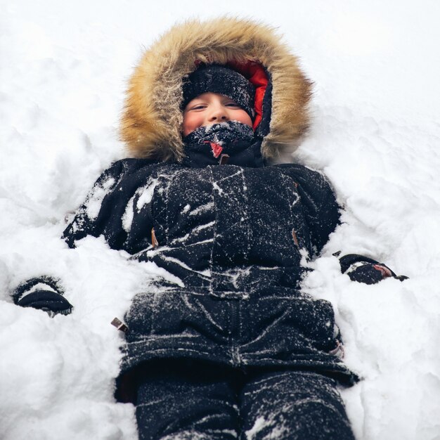 Felice ragazzo vestito con una calda giacca invernale con cappuccio di pelliccia giace nella neve Divertimento invernale