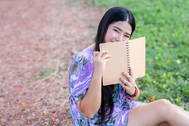 Felice rilassante Ritratto di donna asiatica freelance Indossare un abito viola mentre si lavora tenendo un diario per scrivere una nota mentre si siede sul prato verde dell'erba accanto a un serbatoio nel parco cittadino all'aperto