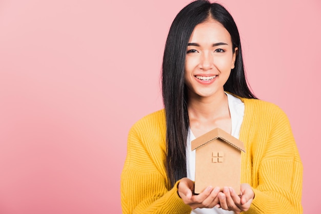 Felice ritratto asiatico bella giovane donna carina eccitata sorridente tenendo il modello di casa a portata di mano, girato in studio isolato su sfondo rosa, broker femminile tenere casa assicurazione immobiliare e concetto bancario