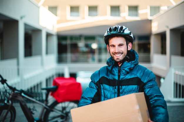 Felice ritratto del corriere con il pacco da consegnare. Sfondo di biciclette. Consegna a domicilio, trasportatore.