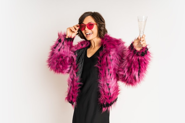 Felice selebrating donna in elegante pelliccia che tiene in mano un bicchiere di champagne che indossa una pelliccia rosa e posa su bianco.