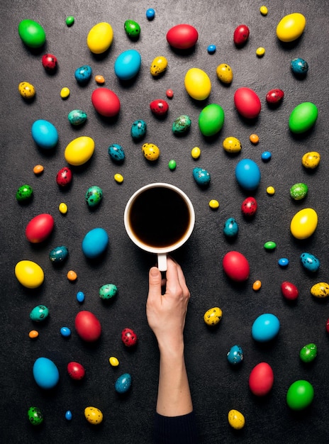 Felice sfondo di Pasqua Una tazza di caffè in una mano di una donna su uno sfondo nero di uova di Pasqua colorate decorate e cioccolato Piatto creativo di instagram Vista dall'alto