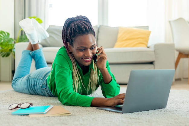 Felice signora afroamericana sdraiata sul pavimento e parlando al cellulare utilizzando il computer portatile che lavora da casa