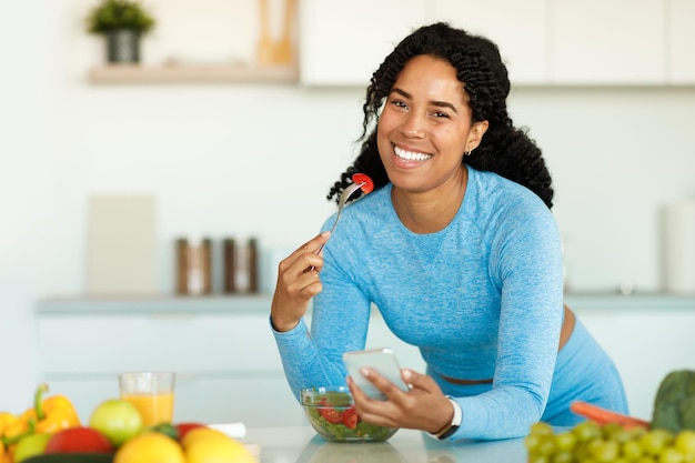 Felice signora nera in abiti sportivi che mangia insalata a casa in cucina appoggiata sul tavolo e utilizzando lo spazio libero dello smartphone