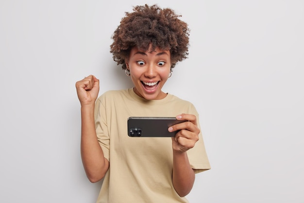 Felice sorpresa donna dai capelli ricci celebra la vittoria del videogioco su smartphone fissa sorprendentemente alla fotocamera finalmente superato il livello difficile vestito con una maglietta casual isolato su sfondo bianco