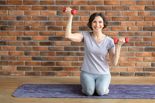 Felice sorridente donna anziana praticante di fitness, seduto sul tappetino fitness con manubri a casa