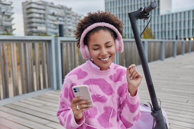 Felice sorridente donna dai capelli ricci in maglione casual tiene smartphone moderno gode di ascoltare musica tramite le cuffie esprime emozioni sincere pone vicino scooter elettrico utilizza il trasporto ecologico in città