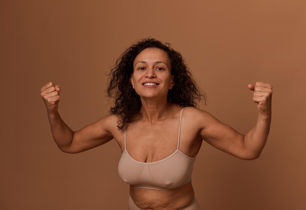 Felice sorridente donna riccia dai capelli scuri in biancheria intima beige, stringendo i pugni, sorridendo con un sorriso a trentadue denti, guardando la telecamera. Positività corporea, concetto di lotta all'oppressione dei diritti delle donne