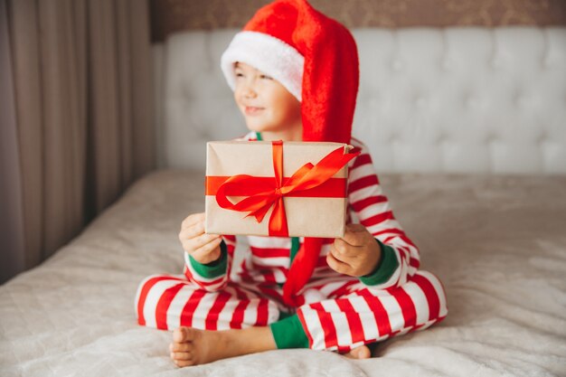 Felice sorridente gioioso bambino con cappello di Babbo Natale, con in mano un regalo di Natale, seduto sul letto.