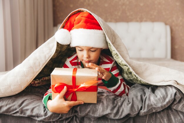 Felice sorridente gioioso bambino con cappello di Babbo Natale, con in mano un regalo di Natale, seduto sul letto.