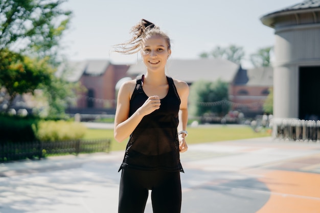 Felice sportiva energica corre allo stadio ha una coda di cavallo che galleggia sul vento vestita con abbigliamento attivo essere un corridore attivo ha un sorriso positivo gode di regolari allenamenti mattutini ti motiva per lo sport