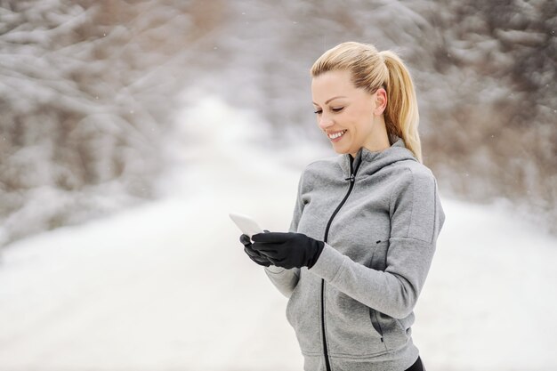 Felice sportiva in piedi nella natura al giorno d'inverno nevoso e usando il suo telefono per mandare SMS. Tecnologia, telecomunicazioni, fitness invernale