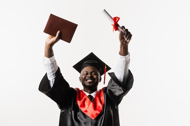 Felice studente africano in abito da laurea nero e berretto solleva il diploma di laurea magistrale sopra la testa su sfondo bianco L'uomo africano laureato si sta laureando e celebrando i risultati accademici