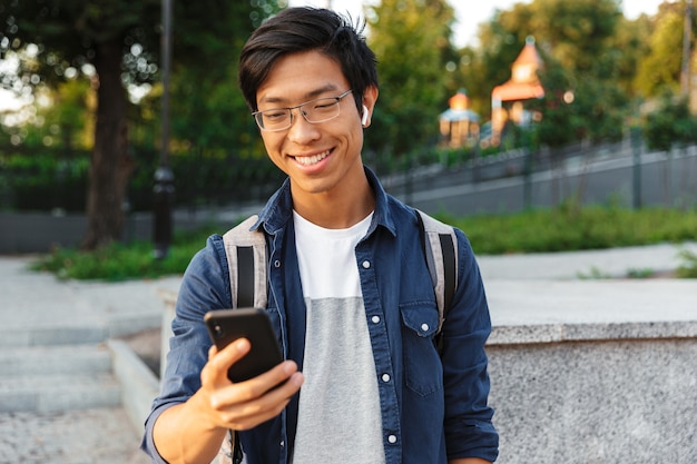 Felice studente maschio asiatico in occhiali e auricolari utilizza lo smartphone all'aperto