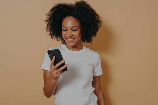 Felice studentessa africana sorridente che utilizza il telefono cellulare e sorride, legge notizie positive in internet o chatta con il fidanzato online, tiene in mano uno smartphone moderno mentre posa contro il muro color sabbia