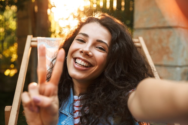 Felice studentessa allegra e mostrando il segno di pace durante l'assunzione di foto selfie mentre è seduto in poltrona nel parco in giornata di sole