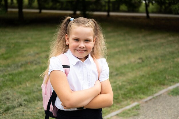 Felice studentessa bionda riccia in uniforme scolastica con zaino rosa torna a scuola all'aperto