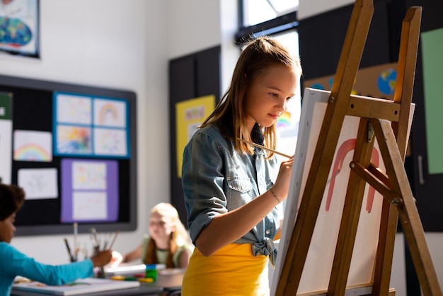 Felice studentessa caucasica che dipinge usando pennello e cavalletto nella classe d'arte della scuola