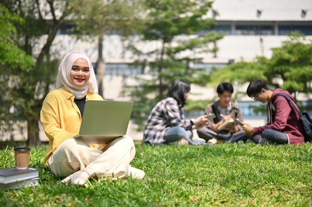 Felice studentessa universitaria musulmana asiatica in hijab seduta sull'erba e usando il suo computer portatile