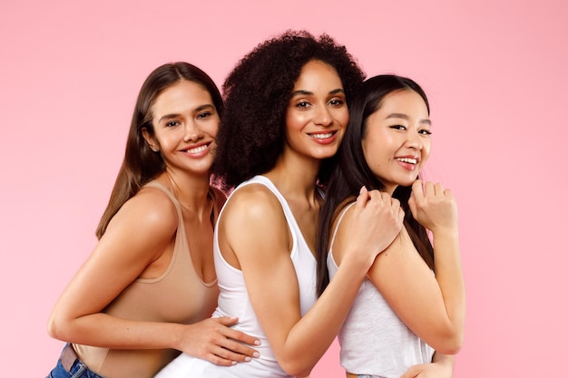 Felice tre donne diverse che posano e abbracciano in piedi su sfondo rosa studio e sorridono a