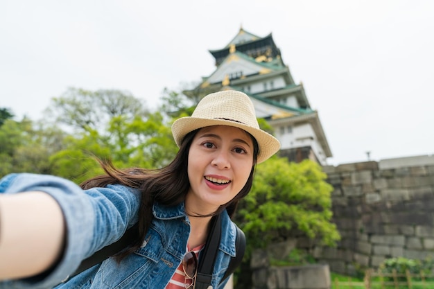 felice turista asiatico ragazza che indossa a prendere autoritratto con il castello di Osaka in giappone in primavera. sorride alla telecamera con l'attrazione storica sullo sfondo