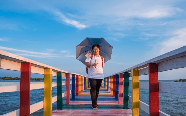 Felice turista femminile asiatico che tiene ombrello e posa sul ponte di legno colorato al punto di vista del mare