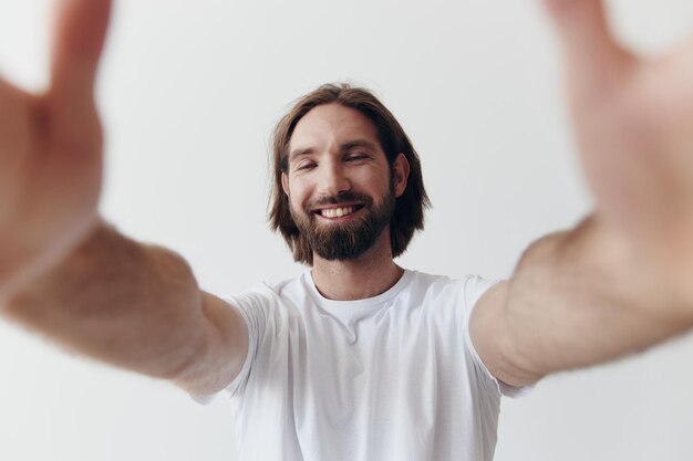Felice uomo adulto con la barba sorride e tira il cuoco nella fotocamera ascoltando musica in cuffia in una maglietta angosciata su uno sfondo bianco isolato