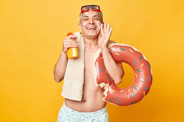 Felice uomo adulto divertente che indossa pantaloncini costume da bagno occhiali da snorkeling tenendo anello di gomma ciambella e borsa isolata su sfondo giallo applicando urlo dal sole che protegge la pelle