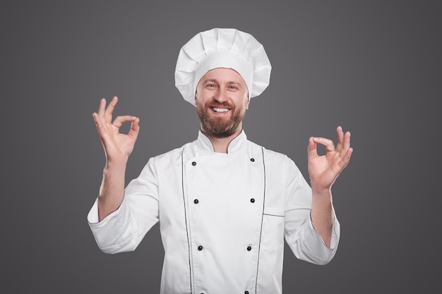 Felice uomo adulto in uniforme da chef che guarda la telecamera con un sorriso felice e mostra un gesto ok su sfondo grigio