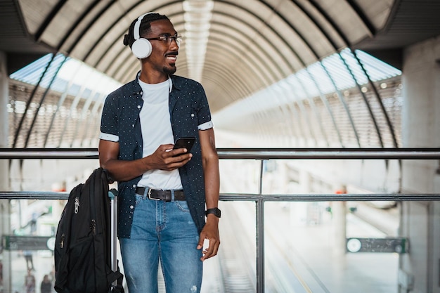 Felice uomo africano che ascolta la musica con le cuffie wireless mentre sta aspettando in una stazione ferroviaria