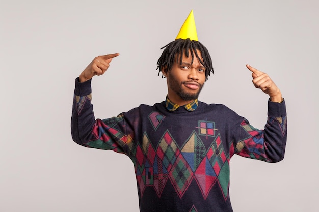 Felice uomo africano soddisfatto con i dreadlocks alla moda che puntano le dita sul cappello da festa sulla sua testa festa festa celebrazione buon umore studio indoor girato isolato su sfondo grigio