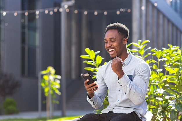 Felice uomo afroamericano in abiti casual seduto sulla panchina all'aperto con il cellulare in mano leggendo buone notizie, vincendo la vittoria guardando lo smartphone esulta fuori. Ragazzo emozioni studente risultato