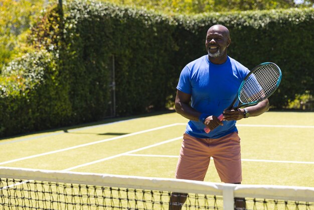 Felice uomo afroamericano senior che gioca a tennis sul campo in erba soleggiata, spazio per fotocopie. Stile di vita senior, pensione, sport, estate, fitness, hobby e attività ricreative.