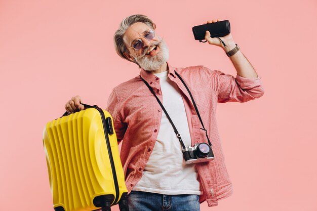 Felice uomo alla moda con la barba in occhiali da sole con la macchina fotografica che tiene una valigia e un altoparlante portatile