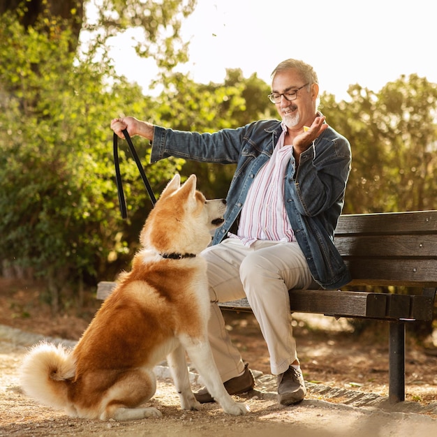 Felice uomo anziano caucasico con barba e occhiali dà uno spuntino al cane si diverte ad allenarsi nel parco